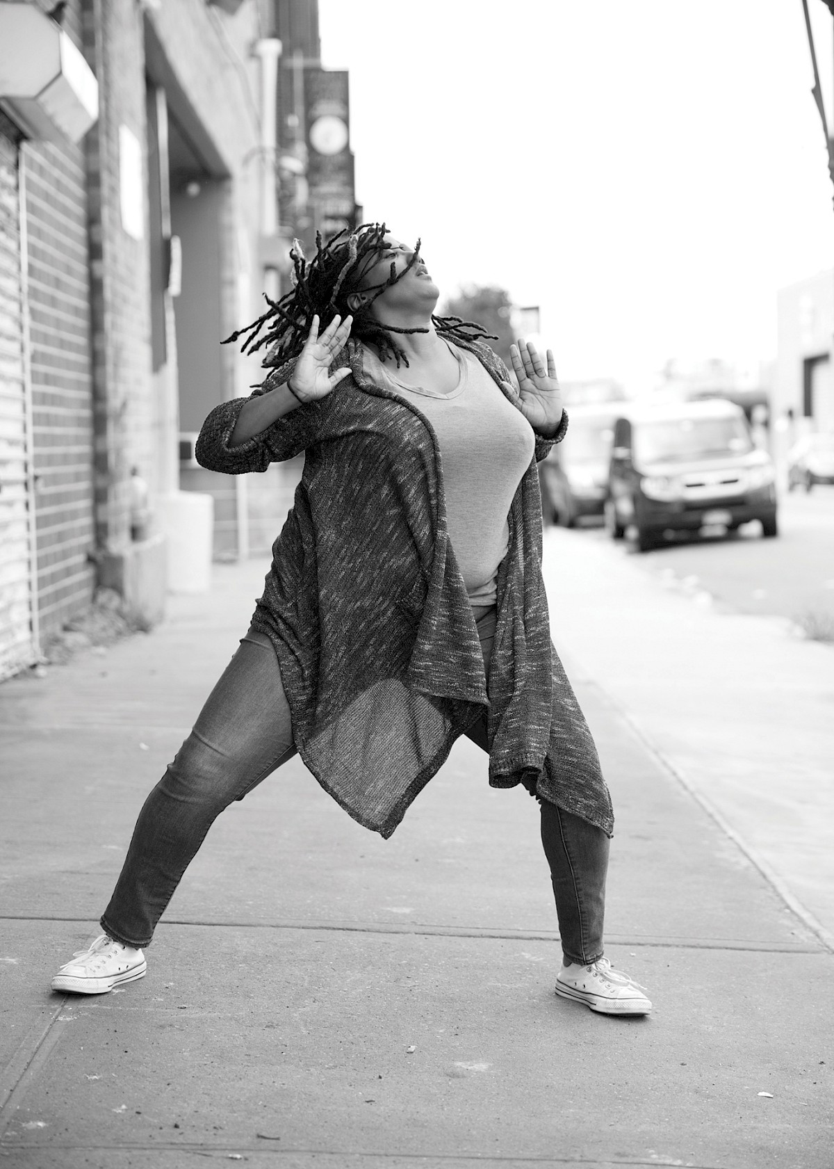 Movement 3—A black and white dance image of Kayla Hamilton, a Black woman, on a city street. She is throwing her head back as her dreads flow with her, and she pushes her arms outward. Her legs are wide and slightly bent. She is wearing jeans and a knee-length cardigan that wraps around her thighs. photo by Travis McGee