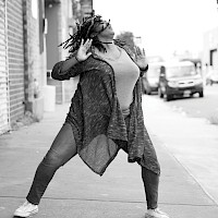 Movement 3—A black and white dance image of Kayla Hamilton, a Black woman, on a city street. She is throwing her head back as her dreads flow with her, and she pushes her arms outward. Her legs are wide and slightly bent. She is wearing jeans and a knee-length cardigan that wraps around her thighs. photo by Travis McGee